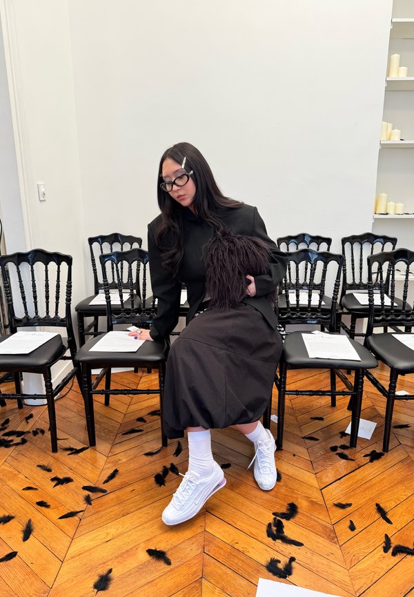 Ženska oblečena v črno sedi na stolu v vrsti črnih stolov, s črnimi peresi raztresenimi po lesenem tleh.