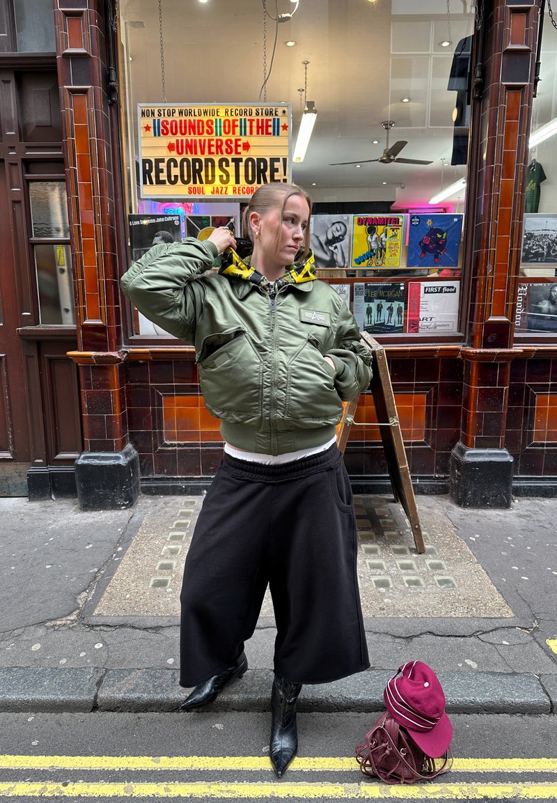 Persona con chaqueta verde y pantalones anchos negros de pie en la calle frente a una tienda de discos vintage con portadas de álbumes coloridas exhibidas.