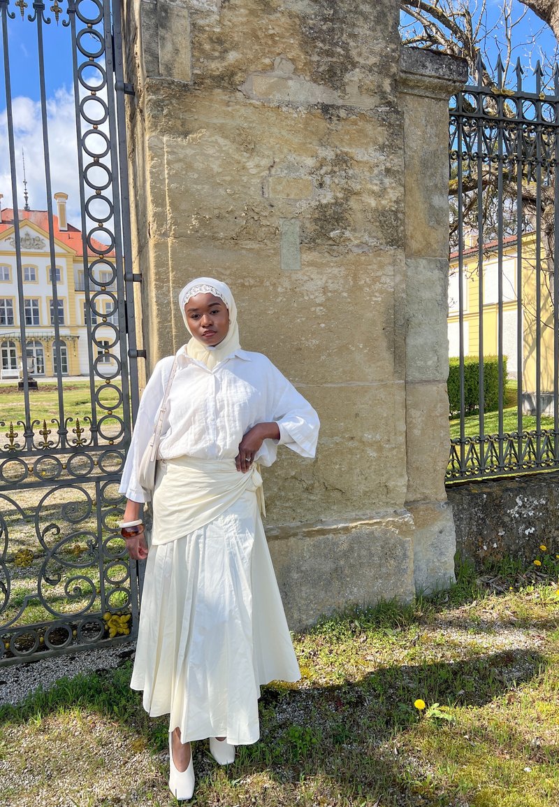 Femme en robe blanche et foulard sur la tête, debout près d’une porte en fer forgé ornée et un mur de pierre sous un ciel bleu clair.