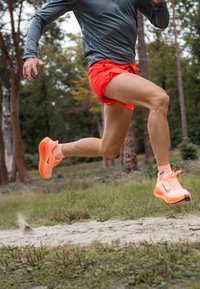 Atleta w szarej bluzie z długim rękawem i pomarańczowych szortach biega po ściśniętej ścieżce, nosząc jasnopomarańczowe i szare sneakersy z widocznym logo marki.