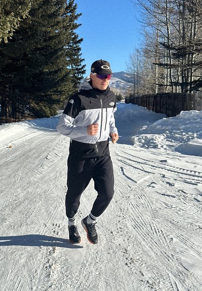 Atleta corriendo por un camino cubierto de nieve, vistiendo una chaqueta cortavientos blanca y negra, pantalones negros y zapatillas deportivas. Fondo soleado.