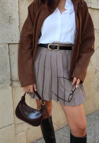 Veste en suède marron, chemise blanche, ceinture noire avec boucle dorée, jupe plissée à carreaux, sac à main brillant marron foncé et bottes noires montantes.