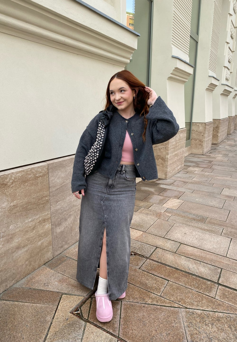 Grå ull cardigan over en rosa topp, denimskjørt med frontsplit, og rosa sko. Bærer en svart veske med sølvnagler. Urban setting.