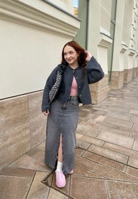 Grå ull cardigan over en rosa topp, denimskjørt med frontsplit, og rosa sko. Bærer en svart veske med sølvnagler. Urban setting.