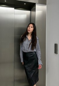 Jupe en cuir noir associée à une chemise rayée noir et blanc. Texture lisse, design ajusté et fermeture à boutons sur la chemise.