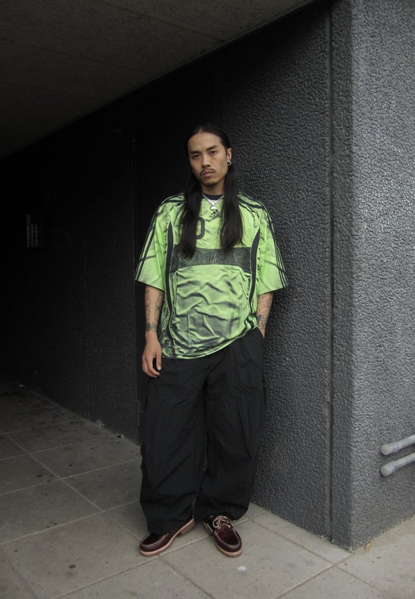 Uomo con capelli lunghi che indossa una camicia verde brillante oversize, pantaloni neri larghi e scarpe marroni in piedi contro un muro grigio texturizzato all'aperto.