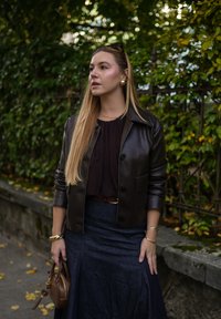 Hnedá kožená bunda cez tmavý blúzku, skombinovaná s džínsovou sukňou. Doplnky zahŕňajú náramok a malú hnedú kabelku.