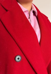 Primo piano di una persona che indossa un cappotto rosso vivace sopra un maglione con zip rosa chiaro, con particolare attenzione ai dettagli del colletto e dei bottoni.