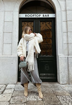 Chaqueta bomber blanca, bufanda blanca con flecos, pantalones de pierna ancha grises, botas vaqueras beige y un bolso acolchado verde frente a una puerta.