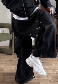 Pantalon noir surdimensionné avec détails à œillets, associé à des baskets blanches. Le tissu a une texture lisse, et le pantalon présente un design en couches.