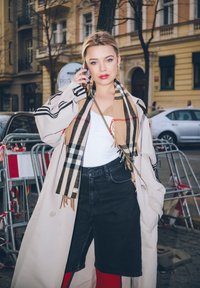 Béžový dlouhý kabát, černé denimové šortky, bílý top a šátek s Burberry vzorem. Drží mobilní telefon, má modré nehty a náušnice ve tvaru kruhu.