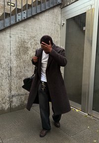 Dark brown double-breasted coat, white T-shirt, black pants, and brown shoes. Carrying a black bag with keys visible on a keyring.