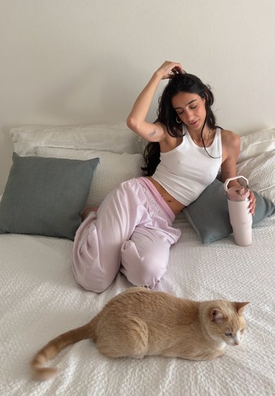 Mujer con camiseta sin mangas blanca y pantalones rosas sentada en la cama sosteniendo una botella de agua, con un gato de color marrón claro acostado en la cama cerca.