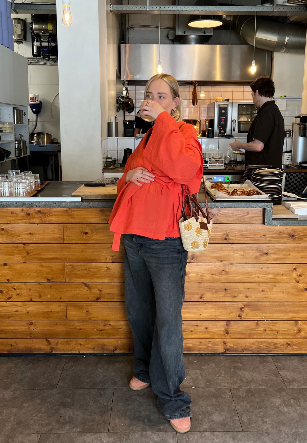 Κόκκινο υπερμεγέθες τοπ τύπου περιτύλιξης, μαύρα παντελόνια με φαρδιά μπατζάκια και ροζ παπούτσια. Υφαντή τσάντα με ανθισμένο σχέδιο. Μπαρ και κουζίνα από ξύλινα πάνελ στο φόντο.