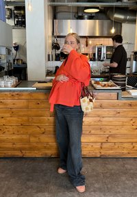 Κόκκινο υπερμεγέθες τοπ τύπου περιτύλιξης, μαύρα παντελόνια με φαρδιά μπατζάκια και ροζ παπούτσια. Υφαντή τσάντα με ανθισμένο σχέδιο. Μπαρ και κουζίνα από ξύλινα πάνελ στο φόντο.