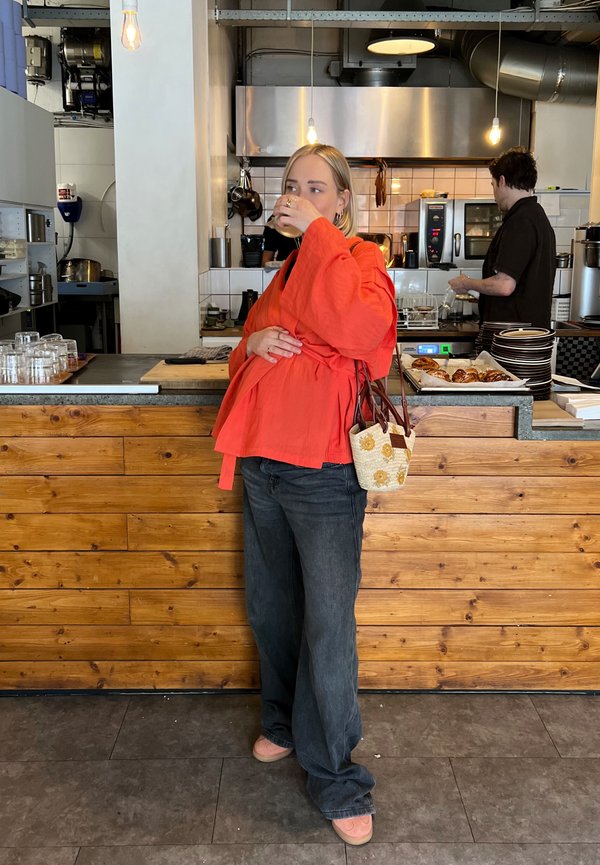 Rode oversized wikkeltop, zwarte wijde jeans en perzikkleurige schoenen. Geweven tas met bloemenpatroon. Houten paneel bar en keuken op de achtergrond.