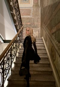 Femme blonde en tenue noire et talons, tenant un sac, montant des escaliers en pierre avec une rampe en fer forgé ornée et des murs peints texturés.