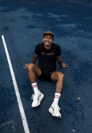 Hombre atlético sentado sobre una superficie azul, vistiendo una camiseta negra TERREX, pantalones cortos negros, zapatillas blancas de Nike y calcetines de marca Nike en blanco.