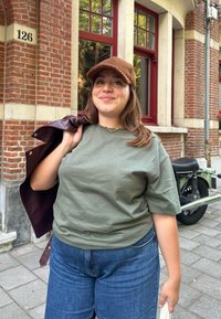 Olivengrøn bomulds t-shirt kombineret med blå denim jeans, holder en burgunder læderjakke. Brun tekstureret hat, murstensvæg baggrund.