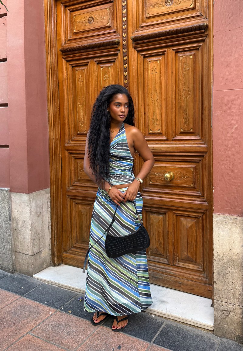 Femme aux longs cheveux bouclés portant une robe dos-nu rayée bleue, verte et blanche, tenant un sac noir, debout près d'une grande porte en bois.