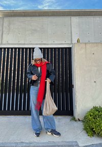 Μαύρο δερμάτινο τζάκετ, κόκκινο φουλάρι με φούντες, μπεζ τσάντα, χαλαρά μπλε τζιν, γκρι καπέλο. Στέκεται μπροστά από έναν τοίχο από σκυρόδε�μα με μαύρες λωρίδες.