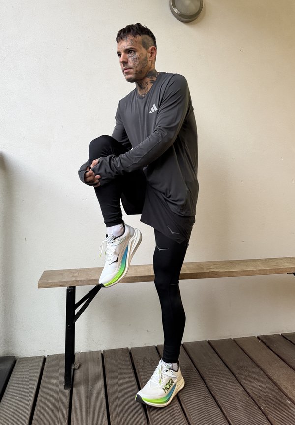 Un hombre tatuado, vestido con ropa deportiva oscura, estira una pierna sobre un banco de madera y lleva zapatillas blancas de running.