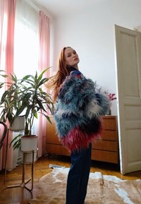 Γυναίκα με μακριά κόκκινα μαλλιά φοράει πολύχρωμο, αφράτο σακάκι, στέκεται σε ένα φωτεινό δωμάτιο με φυτά, ξύλινη τουαλέτα και ροζ κουρτίνες.