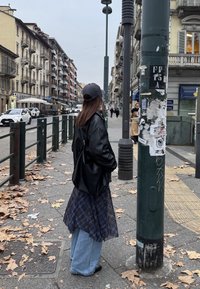 Black leather jacket, long checkered skirt, light blue denim, and black shoes. Standing next to a textured utility pole in an urban setting.