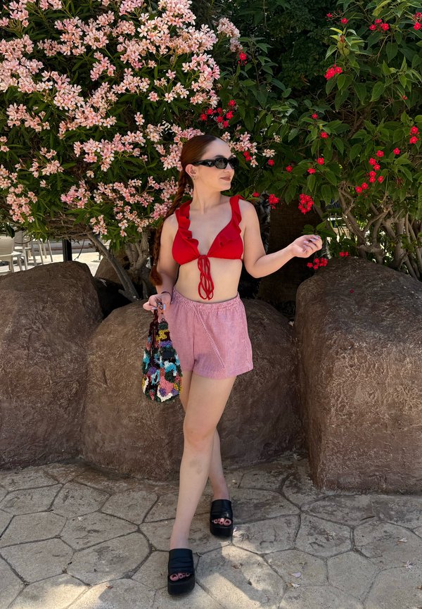 Kvinde iført rød top, pink shorts og sorte sandaler står nær en stor sten og blomstrende buske, mens hun holder en flerfarvet taske.