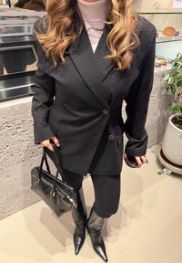 Blazer noir à épaules structurées avec deux boutons, associé à un col roulé clair, des leggings noirs, des bottes montantes et un sac à main noir.