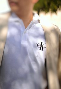Λευκό polo μπλουζάκι με υφή, διακοσμημένο με κουμπί δύο κουμπιών και μαύρο κεντητό λογότυπο που αναγράφει "Calvin Klein Jeans."