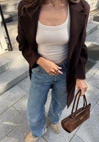 Sacou din lână maro peste un top alb cu caneluri, blugi cu picioare largi de culoare albastră, pantofi slip-on bej și o geantă din ante maro cu două mânere.