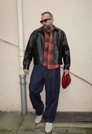 Hombre con gafas de sol que lleva una chaqueta de cuero negra, camisa de cuadros roja, jeans anchos azules, zapatos blancos, sosteniendo una bolsa roja, de pie junto a una pared beige.