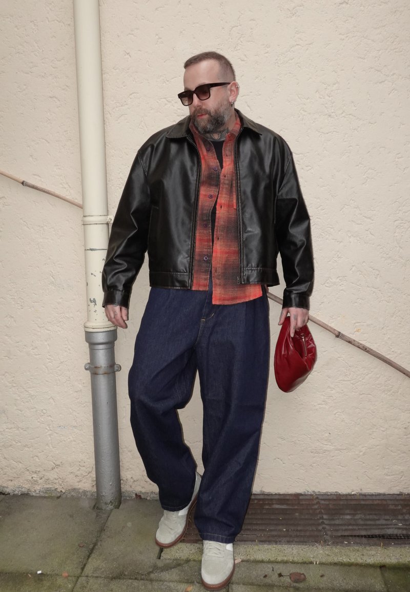 Hombre con gafas de sol que lleva una chaqueta de cuero negra, camisa de cuadros roja, jeans anchos azules, zapatos blancos, sosteniendo una bolsa roja, de pie junto a una pared beige.
