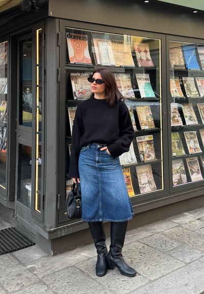 Mujer con suéter negro, falda de mezclilla y botas, parada cerca de una exhibición de revistas afuera de un quiosco en la acera de la ciudad.