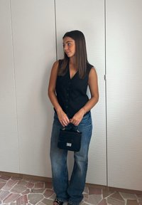 Črn brezrokavnik s gumbi in V-izrezom, v kombinaciji z svetlo modrimi širokimi hlačami. V roki drži majhno črno torbico s kroko.