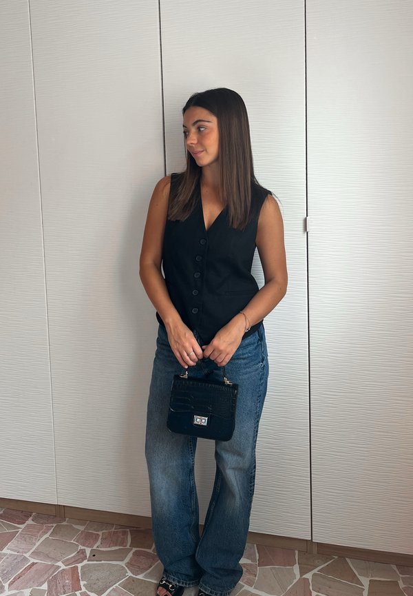 Colete preto sem mangas com decote em V, combinado com jeans de perna larga azul claro. Uma pequena bolsa preta com estampa de croco é segurada.