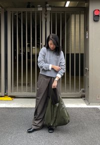 Harmaa neulepusero, jossa on puolivetoketjullinen kaulus, ruskeat leveälahkeiset housut ja kiiltävä oliivinvihreä olkalaukku; seisoo metalliaidaksi taustanaan.