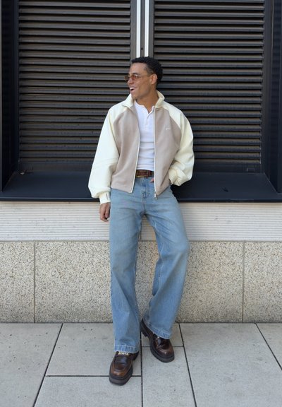 Hombre con gafas de sol, chaqueta con cremallera en crema y beige, camisa blanca, vaqueros azul claro y zapatos marrones, apoyado contra una ventana negra con persianas.