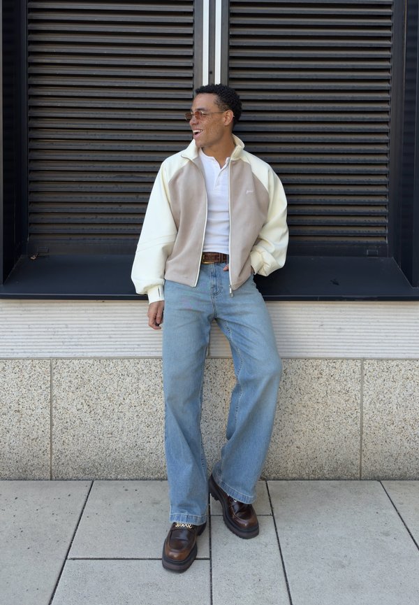 Bărbat purtând ochelari de soare, jachetă crem și bej cu fermoar, cămașă albă, blugi albastru deschis și pantofi maro, sprijinit de o fereastră neagră cu obloane.