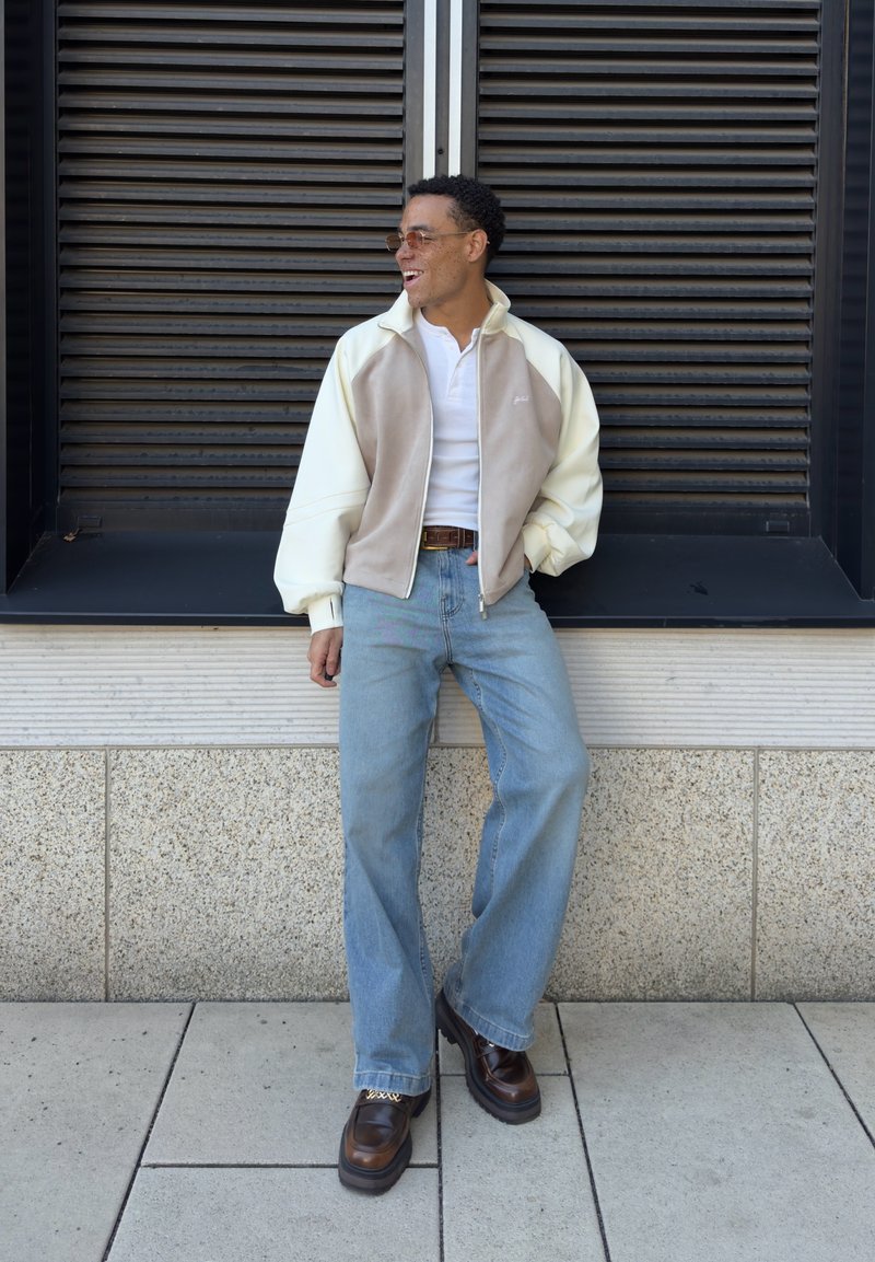 Άντρας με γυαλιά ηλίου, μπουφάν με φερμουάρ σε κρεμ και μπεζ χρώμα, λευκό πουκάμισο, ανοιχτό μπλε τζιν και καφέ παπούτσια, ακουμπώντας σε μαύρο παράθυρο με περσίδες.