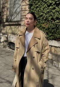 Manteau beige avec un large col et de gros boutons, porté avec un T-shirt blanc et un pantalon noir. Les accessoires comprennent des boucles d'oreilles en or.