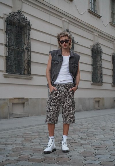 Pantalones cortos de carga con estampado de leopardo, chaleco de mezclilla negro sobre una camiseta blanca sin mangas y botas de combate blancas con cordones en una calle de adoquines.