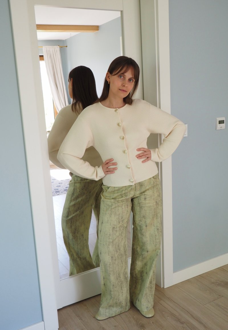 Femme aux cheveux bruns, vêtue d’un pull crème boutonné et d’un pantalon vert ample, se tient les mains sur les hanches devant un miroir en pied.
