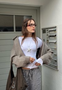 Szary, oversize'owy płaszcz na białej koszuli z długim rękawem zapinanej na guziki, srebrny naszyjnik w kształcie gwiazdy, szare spodnie i ciemnobrązowa torba na ramię.