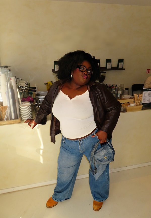 Donna con capelli ricci e occhiali in piedi vicino al bancone del caffè, indossa un top bianco, giacca marrone, jeans larghi, scarpe marroni, tiene una borsa di denim.