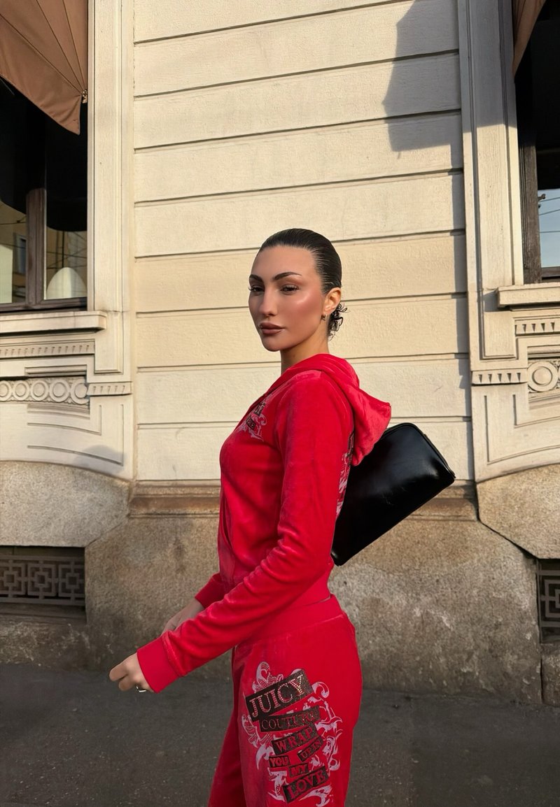 Femme en survêtement Juicy Couture rouge marchant à côté d'un bâtiment de couleur crème, tenant un sac noir sous le bras.