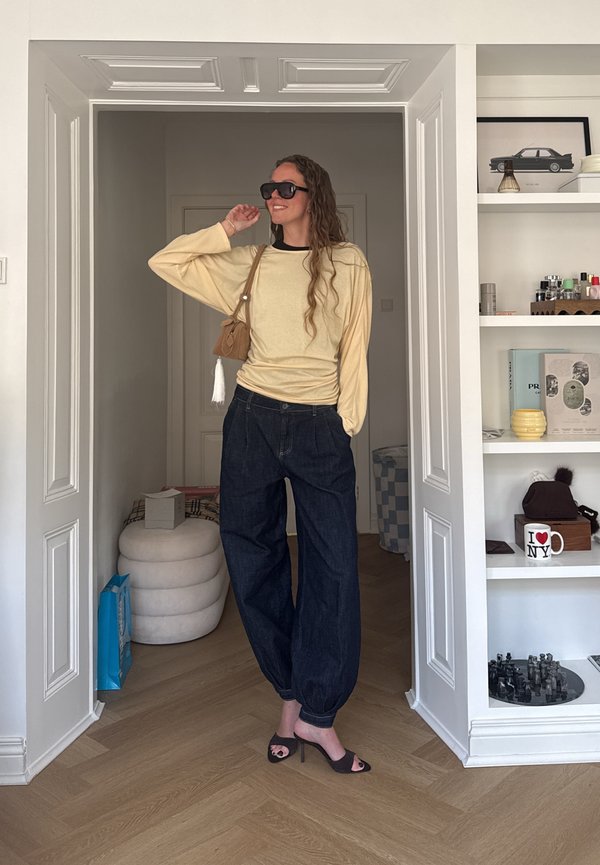 Kobieta w oversize beżowej bluzie, ciemnych szerokich spodniach, czarnych szpilkach, okularach przeciwsłonecznych oraz z małą jasnobrązową torebką na ramieniu pozuje w drzwiach nowoczesnego pomieszczenia.