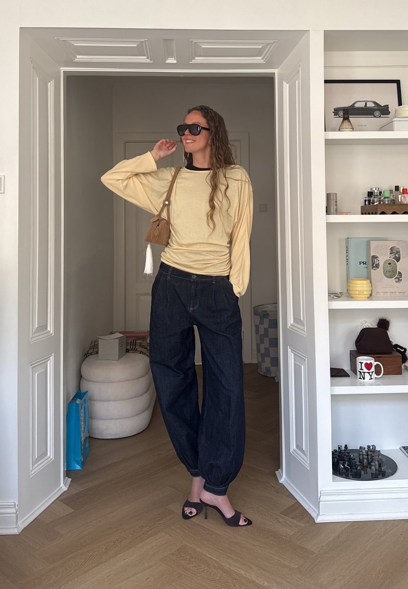 Femme en sweat beige oversize, pantalon large foncé, talons noirs, lunettes de soleil et petit sac bandoulière beige posant dans l’embrasure d’une pièce moderne.