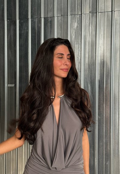 Top gris con drapeados y un acento metálico en el cuello, con tela suave y fluida y detalles fruncidos. El cabello es largo y ondulado.